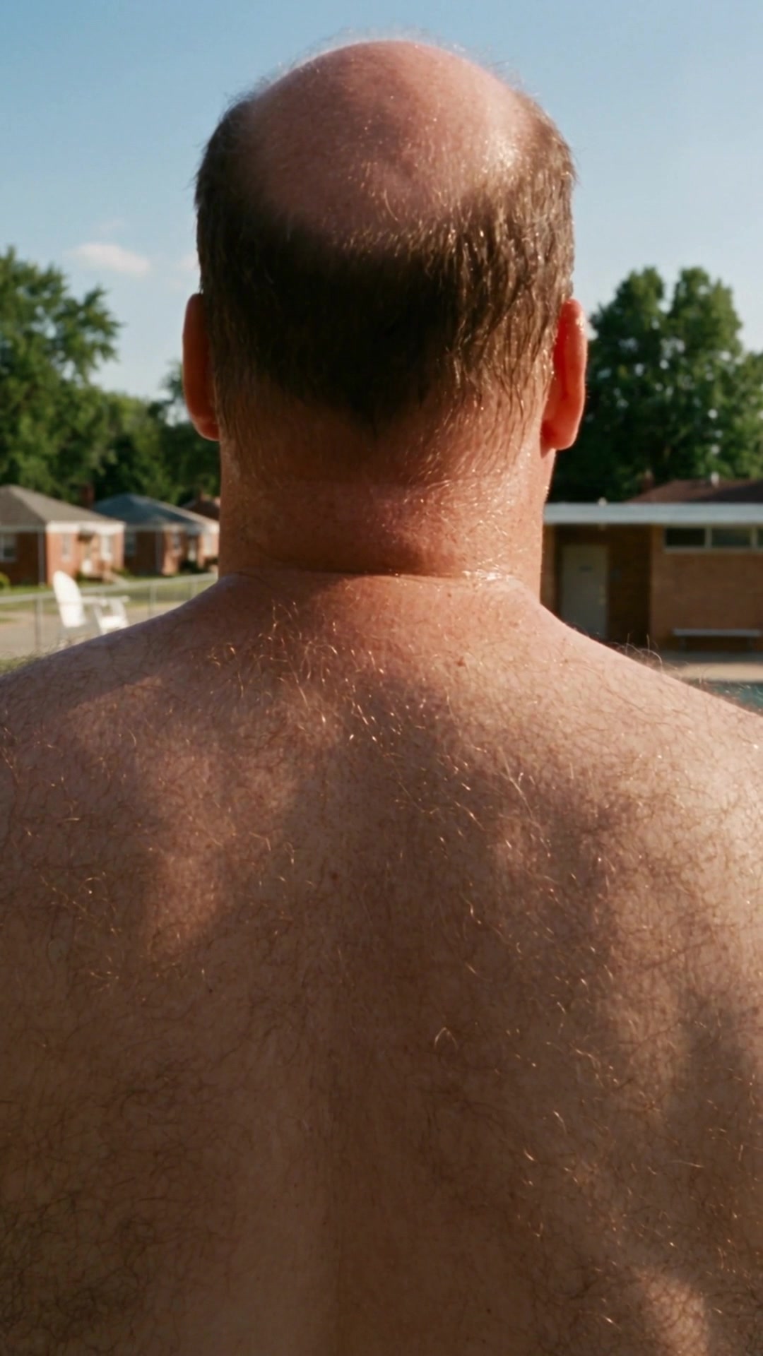 A balding man's bare, hairy back filling the frame at a public pool, the lifeguard chair empty behind him - Ankin Law dramatization