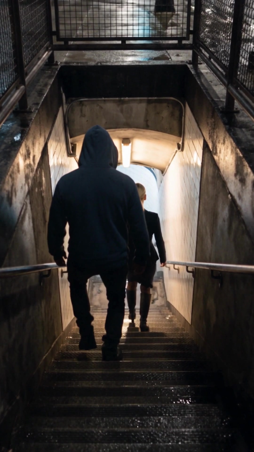 A man in a dark hoodie, seen from behind, following a young woman down the stairs to a subway at night - Ankin Law dramatization