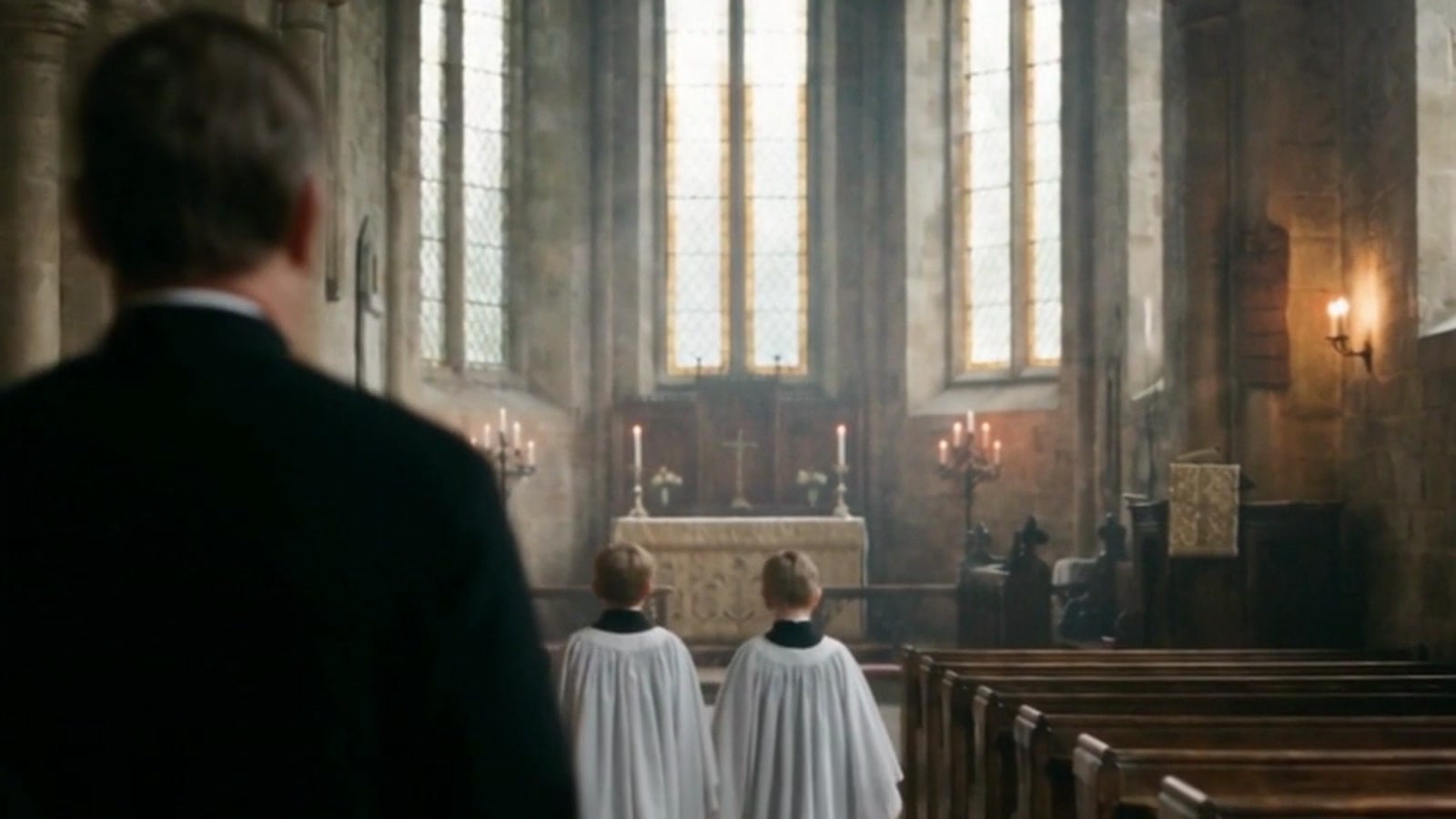 A man in a black cassock, seen from behind, following two altar boys toward the altar