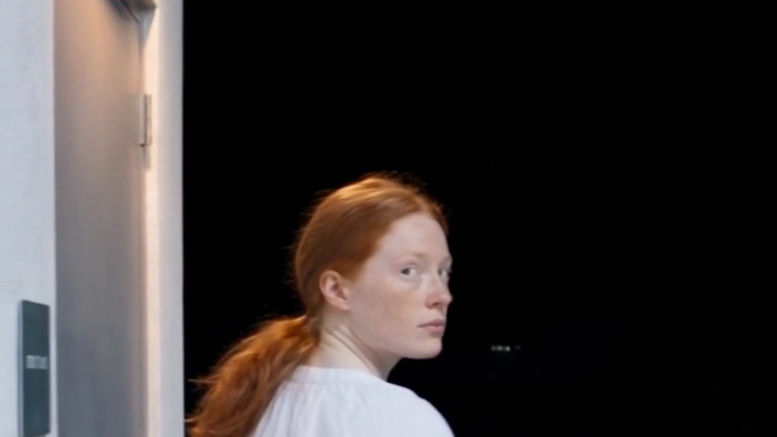A young woman with red hair glancing back over her shoulder at the restroom door of a gas station at night