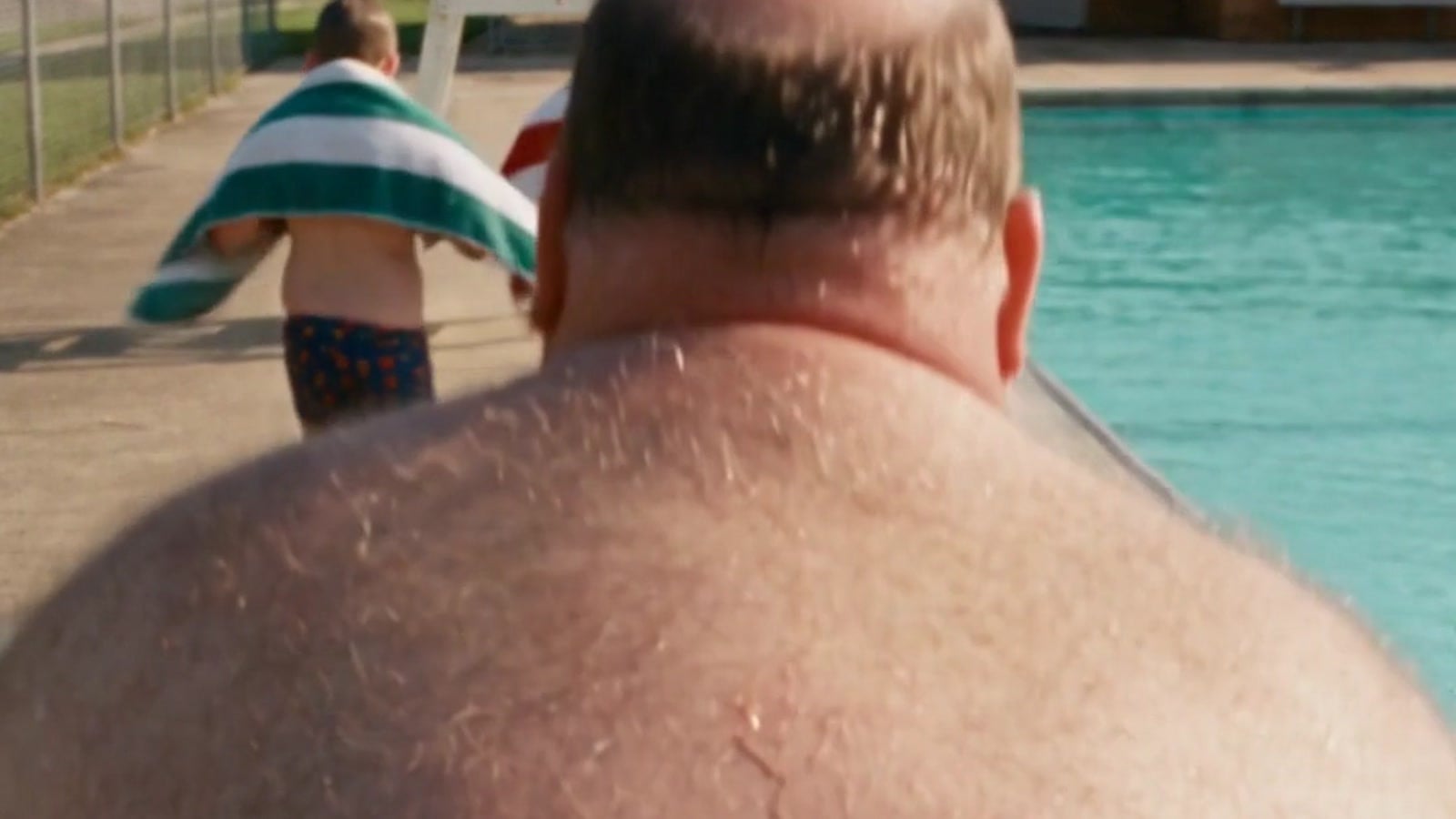 A balding man's bare, hairy back in the foreground as a boy with a towel walks past an empty lifeguard chair