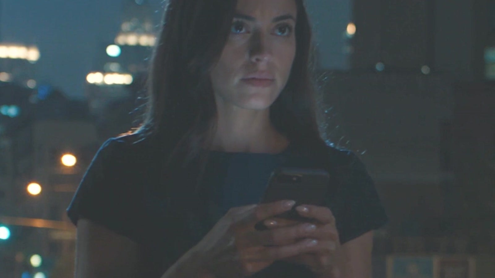 A young woman in a short black dress waiting at the curb at night, looking at her phone