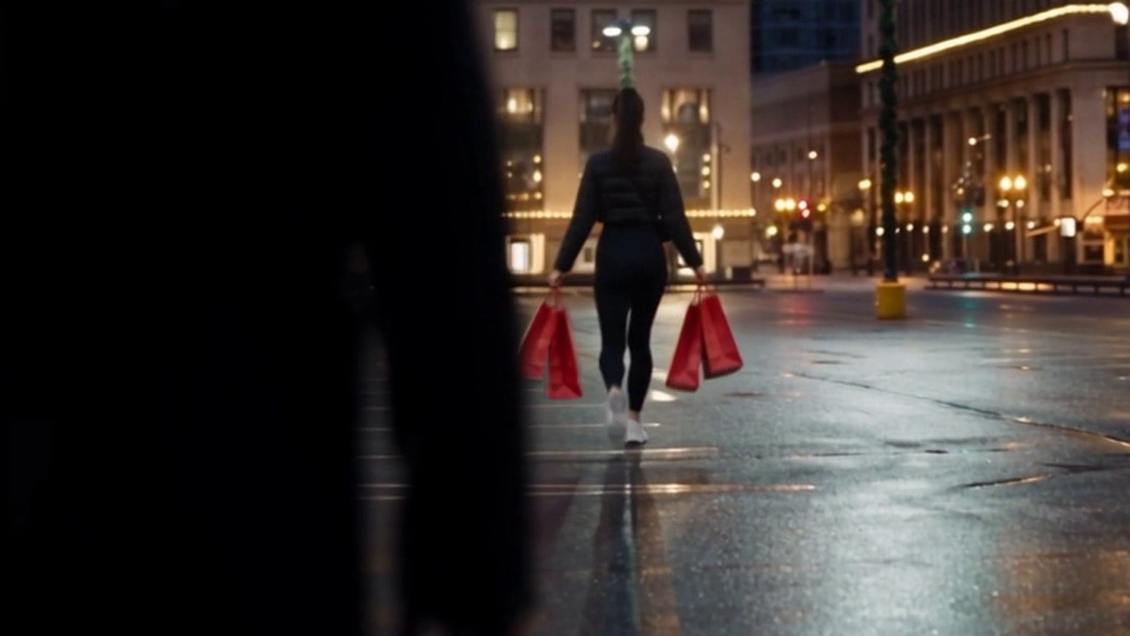 A young woman carrying shopping bags across a dark city parking lot while a figure watches