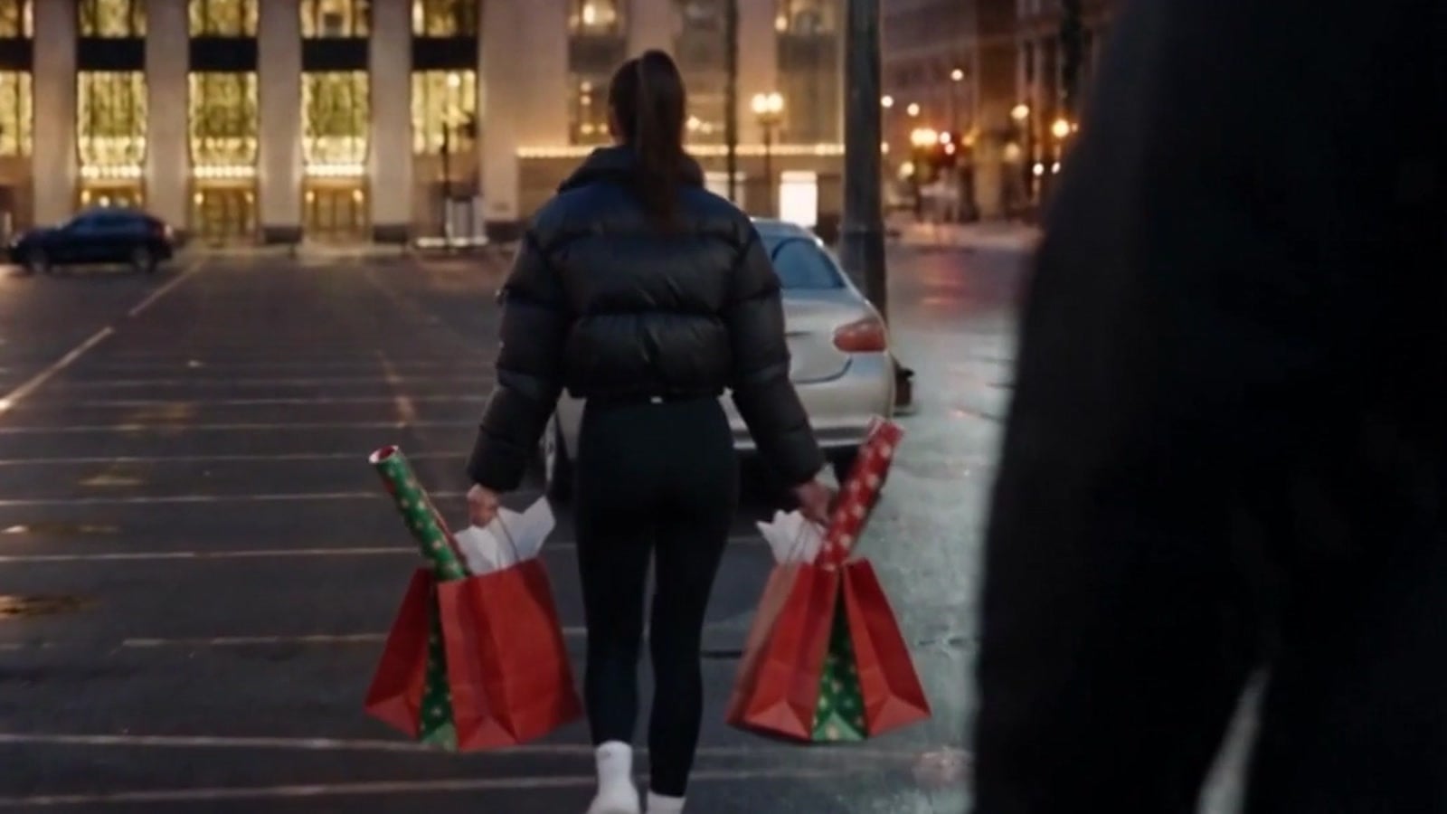 A hooded man's dark silhouette in the foreground as a young woman with shopping bags walks to her car in a city parking lot