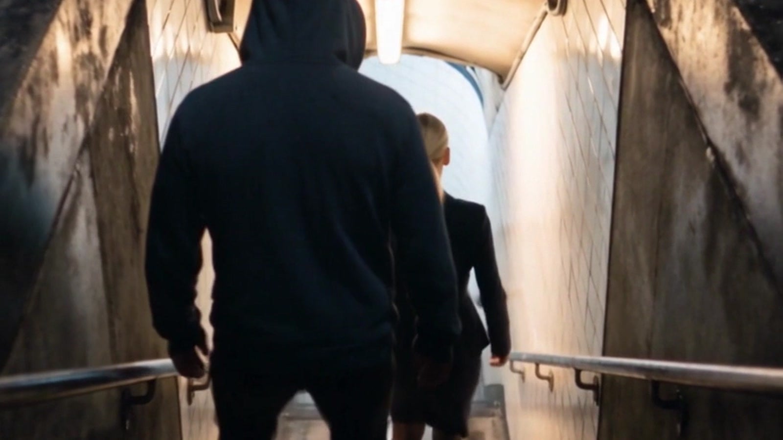 A man in a dark hoodie, seen from behind, following a young woman down the stairs to a subway at night