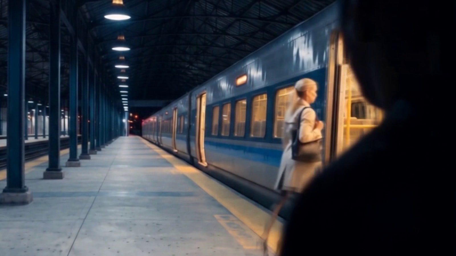 A woman on an empty train platform at night as a figure comes up behind her