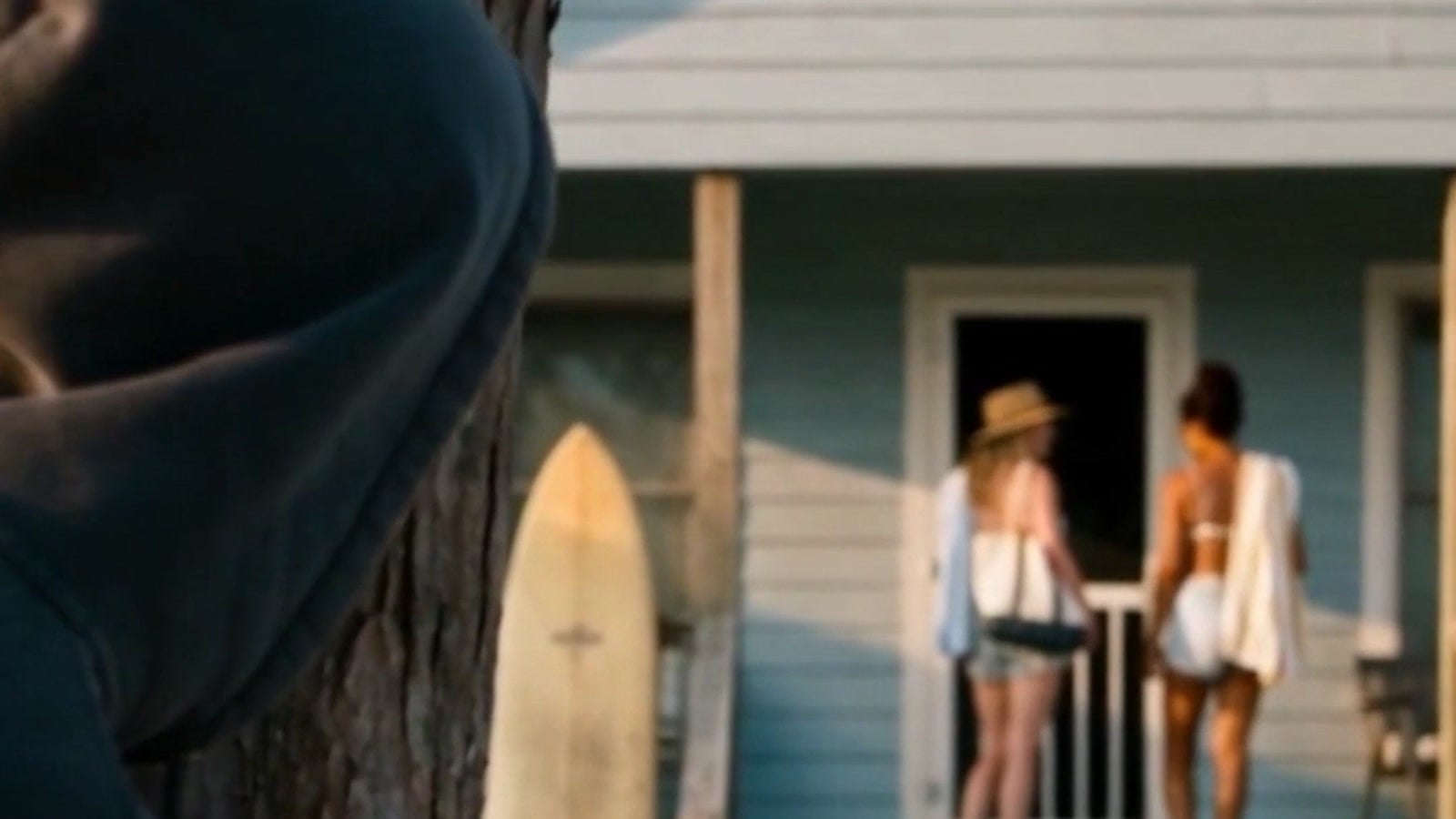 A man in a hood watching from behind a tree as two women stand at the door of a beach vacation rental