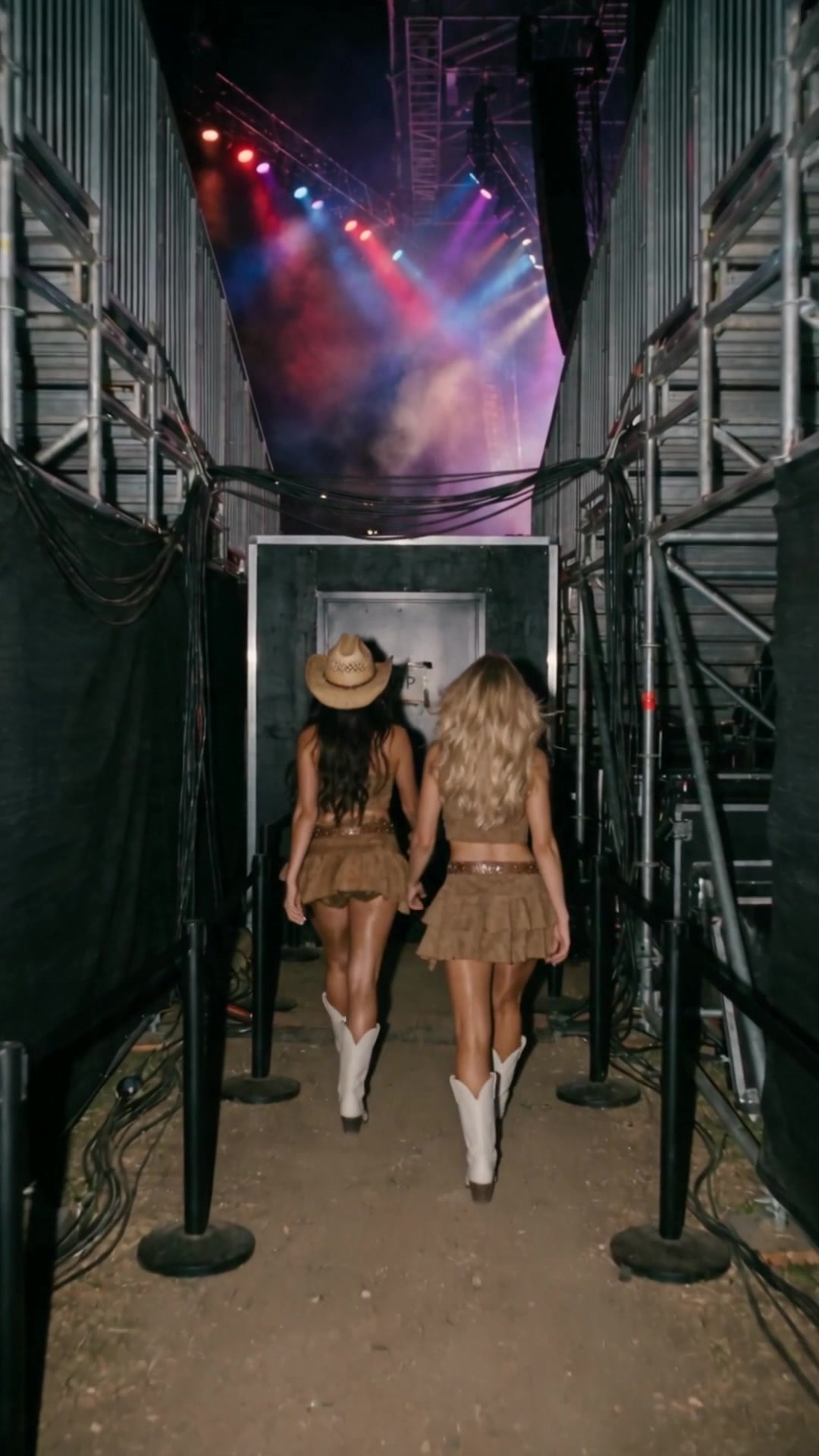 Two young women in cowboy hats and white boots walking down a roped walkway under the stands beside a concert stage - Ankin Law dramatization