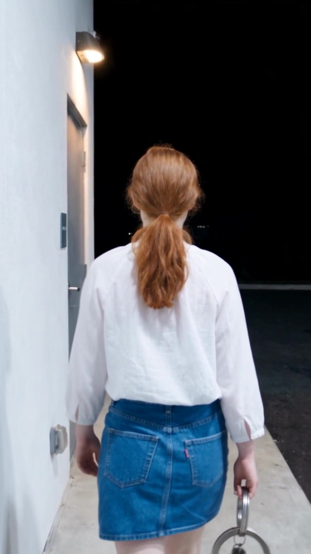 A young woman walking along the white wall of a gas station at night, a restroom key on a big metal ring in her hand - Ankin Law dramatization