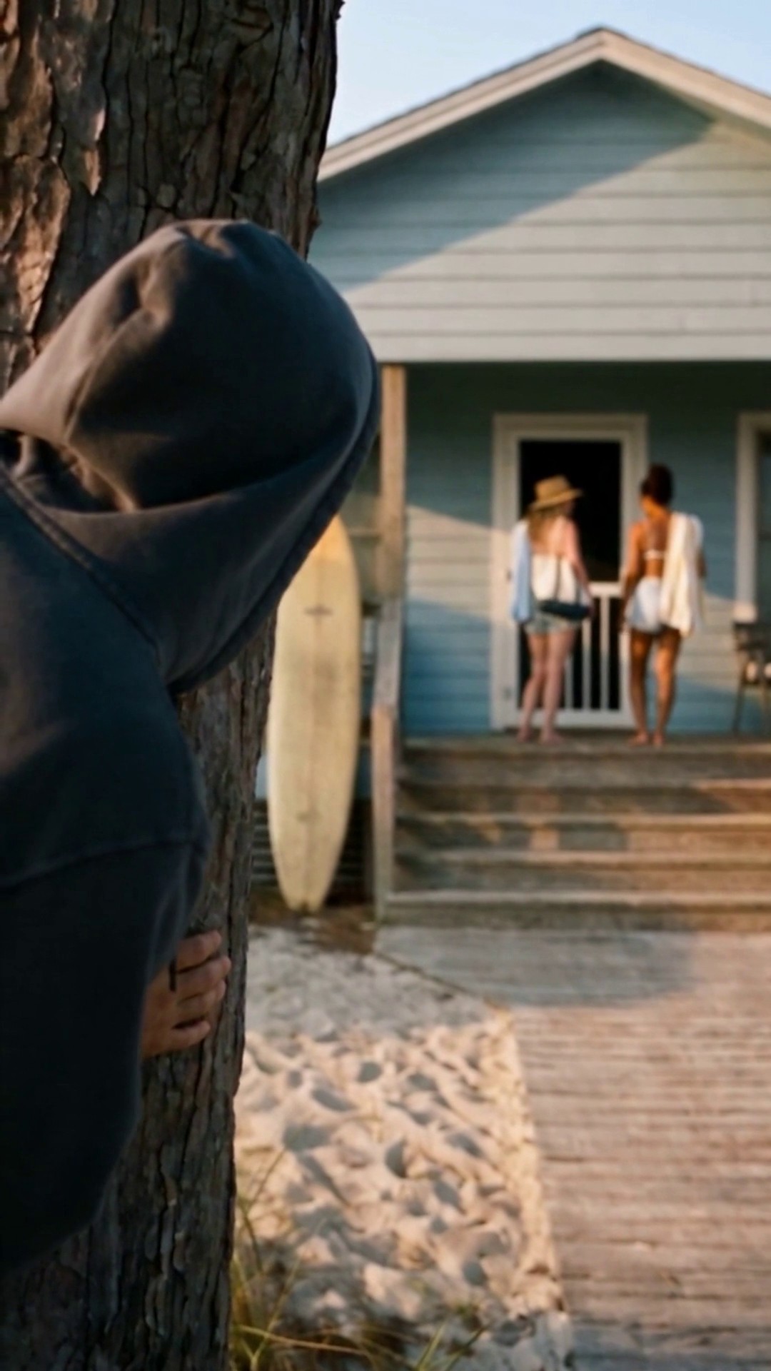 A man in a dark hoodie hiding behind a tree in the foreground, watching two women at the door of a vacation rental - Ankin Law dramatization